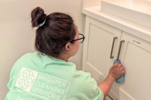 a person in a green brighter cleaning services shirt cleans a bathroom cabinet with a blue cloth. the setting is bright and tidy, conveying professionalism.