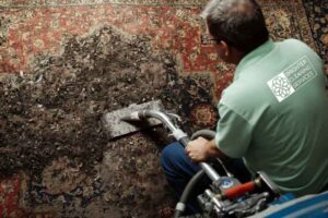 a person in a green shirt vacuums a large, red-and-blue patterned rug covered with dirt and debris. the task appears challenging and focused for carpet cleaning services in vancouver