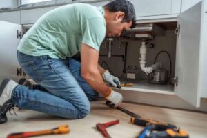 a plumber in a light green shirt and jeans is fixing a pipe under a kitchen sink, surrounded by tools. the scene conveys focus and diligence for handyman services.
