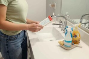 a person in a green shirt and jeans stands by a bathroom sink, holding a cleaning product. several cleaning sprays, brushes are on the counter and a residential cleaning checklist.